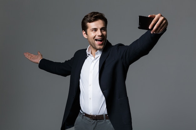 Retrato de un joven empresario emocionado vestido con traje aislado sobre pared gris, tomando un selfie