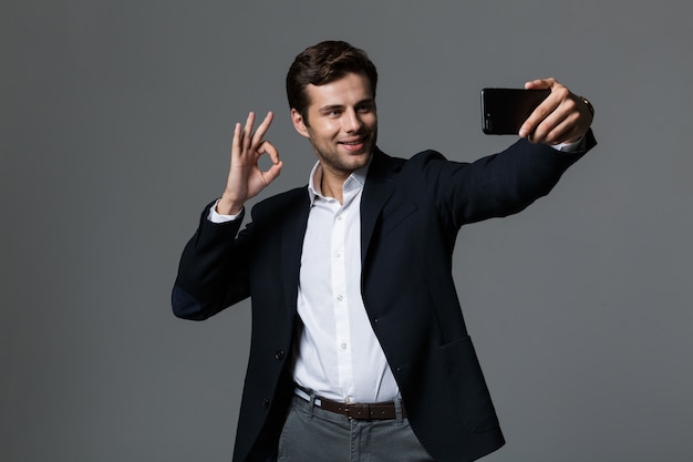 Retrato de un joven empresario feliz vestido con traje aislado sobre pared gris, tomando un selfie