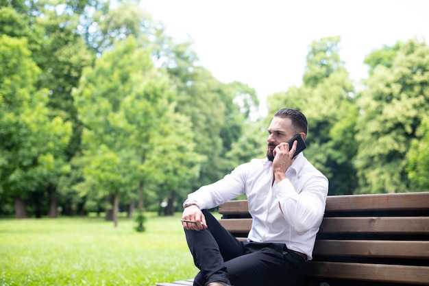 Retrato de un joven empresario físicamente apto fuma cigarro cubano en el parque y habla por teléfono móvil