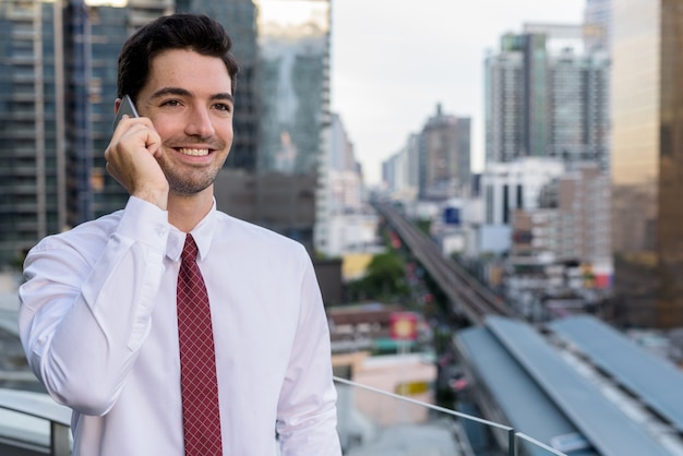 Retrato de joven empresario guapo en la ciudad hablando por teléfono