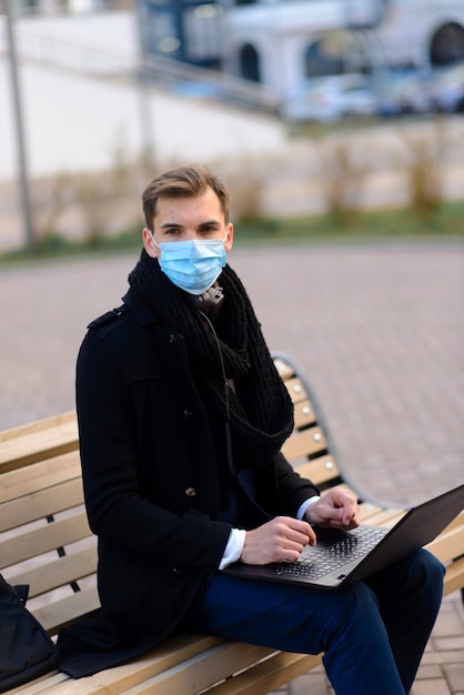 Retrato de joven empresario guapo con mascarilla en cuarentena de gripe. Foto en caffee con laptop, teléfono móvil.