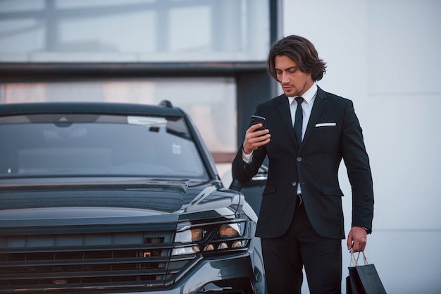 Retrato de joven empresario guapo en traje negro y corbata al aire libre cerca de coche moderno y con bolsas de la compra.