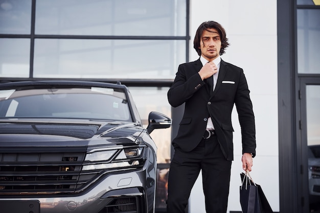 Retrato de joven empresario guapo en traje negro y corbata al aire libre cerca de coche moderno y con bolsas de la compra.