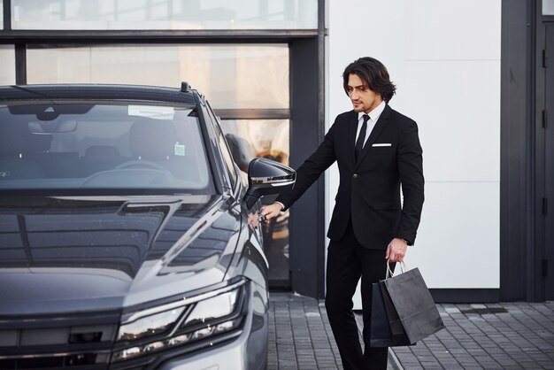 Retrato de joven empresario guapo en traje negro y corbata al aire libre cerca de coche moderno y con bolsas de la compra.