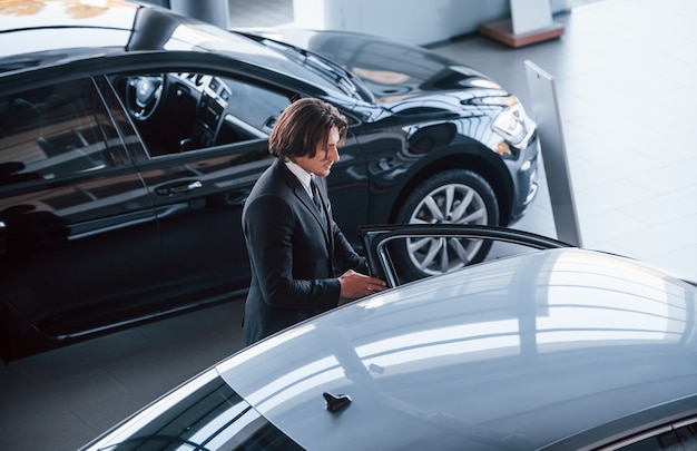 Retrato de joven empresario guapo en traje negro y corbata en el interior cerca de coche moderno.