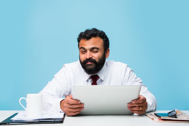 Retrato de joven empresario indio con barba, sentado en una posición relajada en la mesa de la oficina con una mirada agradable. Seguros de hombre de negocios asiático