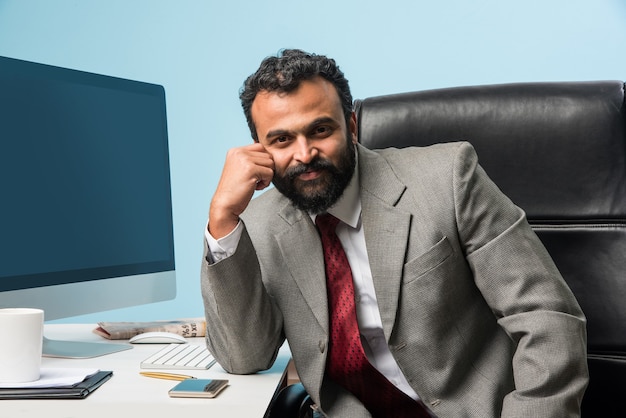 Retrato de joven empresario indio con barba, sentado en una posición relajada en la mesa de la oficina con una mirada agradable. Seguros de hombre de negocios asiático