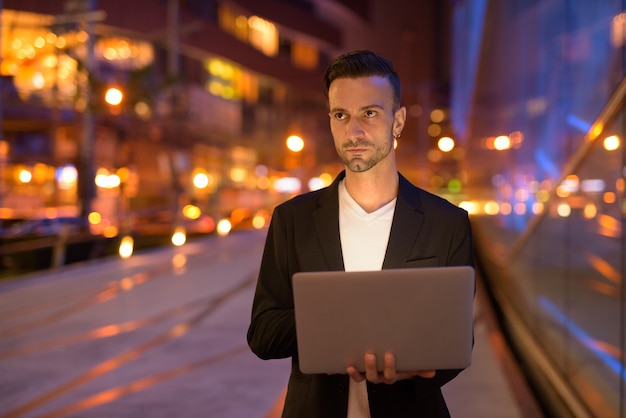 Retrato de joven empresario con portátil contra una ciudad por la noche