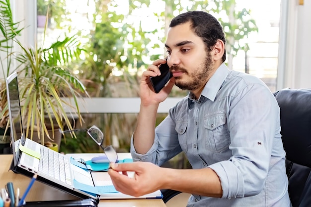 Retrato de un joven empresario sonriente en una oficina informal haciendo una llamada telefónica mientras trabaja con una laptop