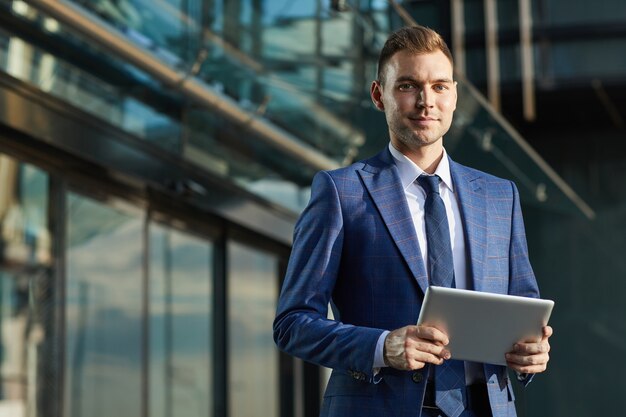 Retrato de joven empresario en traje formal mirando a cámara mientras usa tableta digital en la ciudad