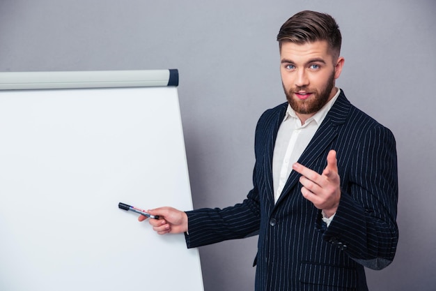 Retrato de un joven empresario en traje presentando algo en tablero en blanco sobre pared gris