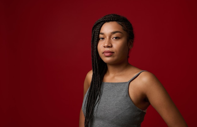 Retrato de una joven y encantadora mujer afroamericana hermosa con elegantes rastas tradicionales, mirando con confianza a la cámara, aislada sobre fondo rojo con espacio de copia