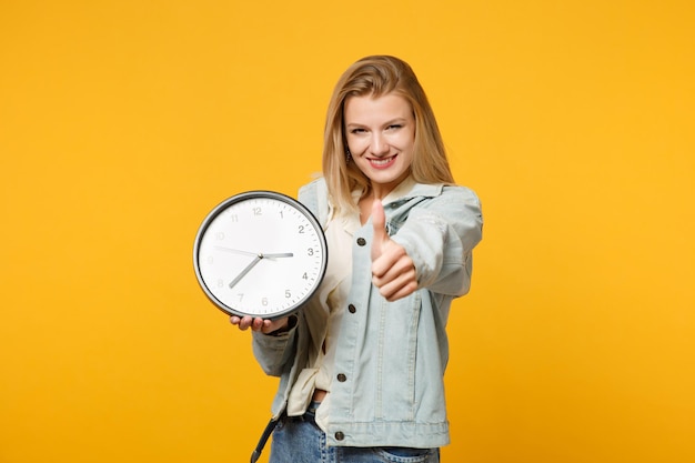 Retrato de una joven encantadora con ropa casual de denim mostrando el pulgar hacia arriba, mirando la cámara, sosteniendo un reloj redondo aislado en el fondo de la pared naranja amarilla. Concepto de estilo de vida de las personas. Simulacros de espacio de copia.