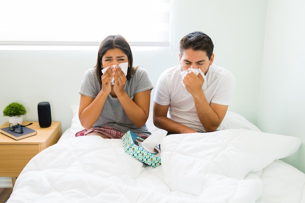 Retrato de una joven enferma y un hombre con gripe sonándose la nariz con papel tisú y sintiéndose mal mientras están acostados juntos en la cama