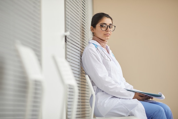 Retrato de joven enfermera en bata blanca mirando al frente mientras está sentado en el pasillo del hospital