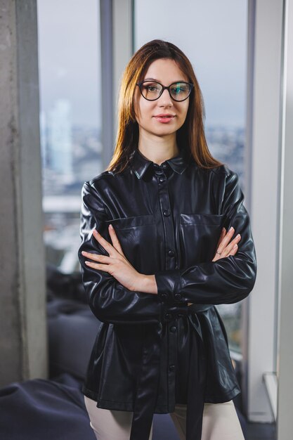 Retrato de una joven esbelta con gafas y una camisa de cuero negro Mujer moderna en el fondo de la ventana de la oficina con una ventana grande