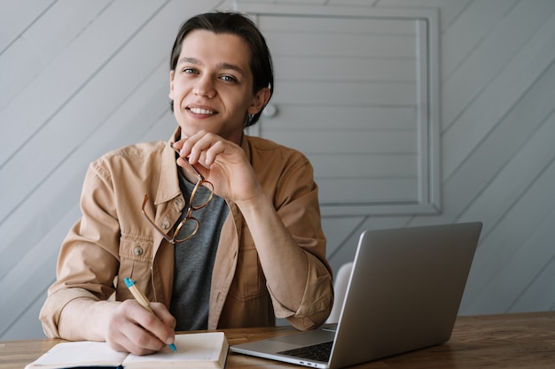 Retrato de joven escritor tomando notas en el cuaderno, con gafas. Freelancer trabajando desde casa