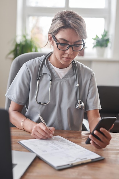 Retrato de una joven especialista médica adulta sentada en el escritorio viendo algo en un teléfono inteligente