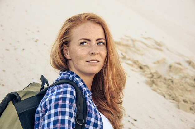 Retrato de joven con estilo hipster con pelo rojo y pecas en una camisa a cuadros en la duna de la pendiente. Con mochila. Concepto de viaje, senderismo y estilo de vida activo.