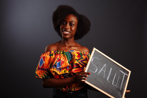 El retrato de una joven estudiante africana vestida sosteniendo una pizarra con una palabra de saludo en su propio idioma nativo aislada de fondo gris