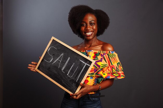 El retrato de una joven estudiante africana vestida sosteniendo una pizarra con una palabra de saludo en su propio idioma nativo aislada de fondo gris