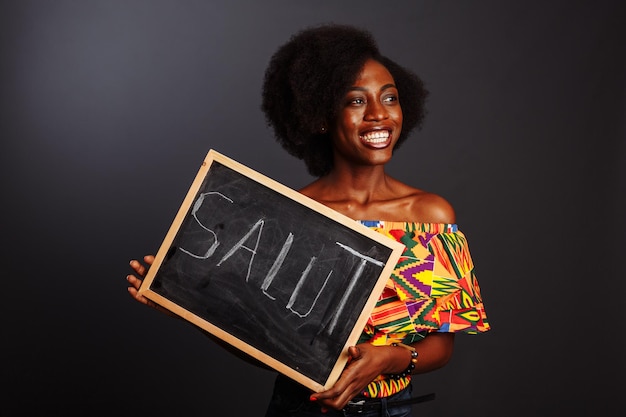 El retrato de una joven estudiante africana vestida sosteniendo una pizarra con una palabra de saludo en su propio idioma nativo aislada de fondo gris