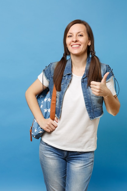 Retrato de joven estudiante alegre bastante feliz en camiseta blanca, ropa de mezclilla con mochila mostrando el pulgar hacia arriba aislado sobre fondo azul. Educación en la universidad. Copie el espacio para publicidad.