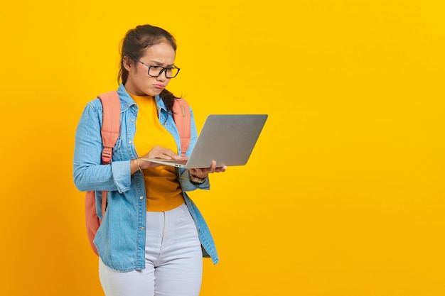 Retrato de una joven estudiante asiática confundida con ropa informal con mochila mirando el correo electrónico entrante en una computadora portátil aislada en un fondo amarillo Educación en el concepto de universidad universitaria