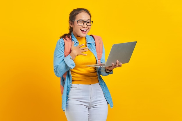 Retrato de una joven estudiante asiática emocionada con ropa informal con mochila usando una computadora portátil con la mano en el pecho aislada en el fondo amarillo Educación en el concepto de universidad universitaria