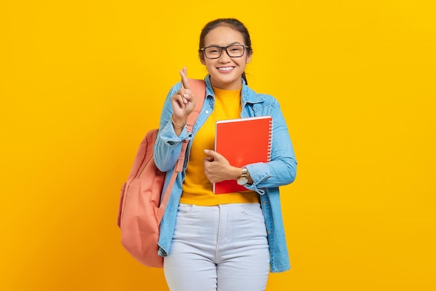 Retrato de una joven estudiante asiática feliz vestida de denim con mochila sosteniendo un cuaderno con los dedos cruzados esperanzados y optimistas para la buena suerte aislada en un fondo amarillo