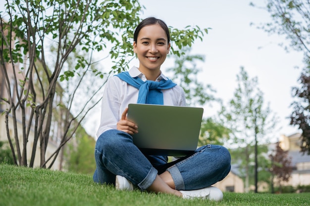 Retrato de joven estudiante asiática hermosa que estudia en línea, sentada sobre la hierba. Concepto de aprendizaje a distancia