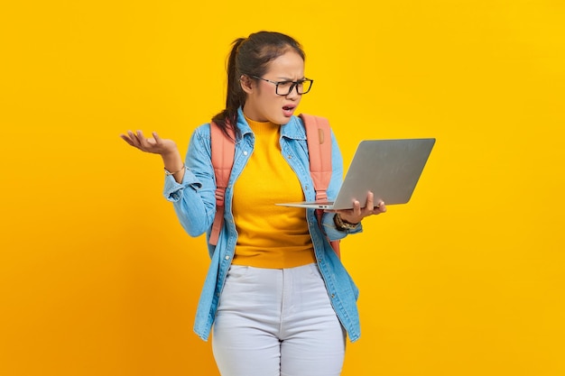 Retrato de una joven estudiante asiática molesta con ropa informal con mochila que muestra espacio de copia en la palma y sostiene una computadora portátil aislada en un fondo amarillo Educación en el concepto de universidad universitaria