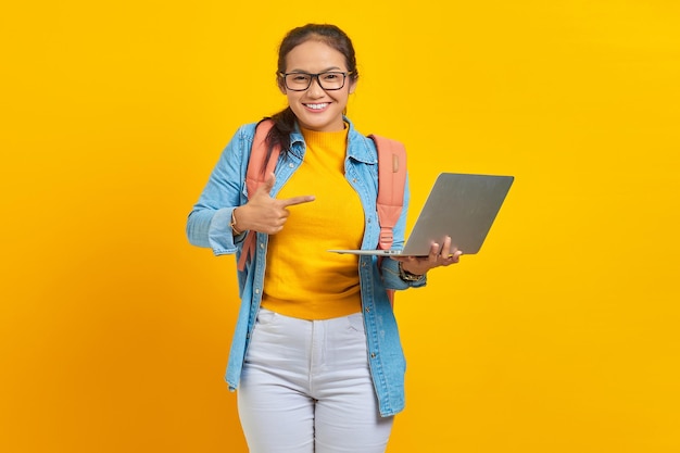 Retrato de una joven estudiante asiática sonriente en ropa casual con mochila apuntando a la computadora portátil con el dedo aislado sobre fondo amarillo Educación en concepto de universidad universitaria