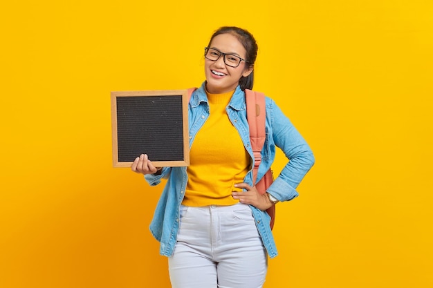 Retrato de una joven estudiante asiática sonriente vestida de forma informal con mochila sosteniendo una pizarra en blanco aislada de fondo amarillo. Educación en concepto de universidad universitaria