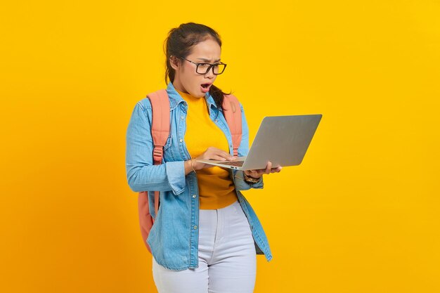 Retrato de una joven estudiante asiática sorprendida con ropa informal con mochila mirando el correo electrónico entrante en una computadora portátil aislada en un fondo amarillo Educación en el concepto de universidad universitaria