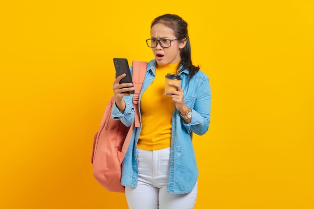 Retrato de una joven estudiante asiática sorprendida vestida de denim con mochila sosteniendo una taza de café y usando un teléfono móvil aislado en un fondo amarillo