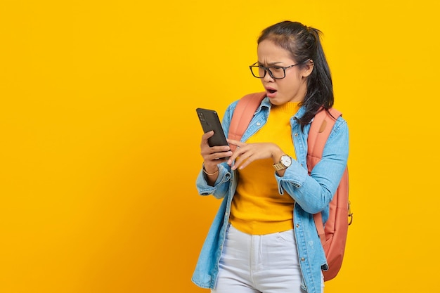 Retrato de una joven estudiante asiática sorprendida vestida de forma informal con mochila escribiendo mensajes de texto o desplazándose en las redes sociales usando un smartphone aislado de fondo amarillo