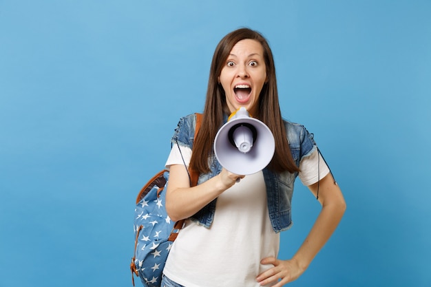 Retrato de joven estudiante encantadora sorprendida en ropa de mezclilla con mochila sosteniendo megáfono electrónico, gritando aislado sobre fondo azul. Educación en la universidad. Copie el espacio para publicidad.