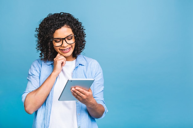 Retrato de joven estudiante estadounidense con cabello africano rizado sosteniendo tableta digital y sonriendo de pie sobre fondo azul aislado con espacio para copiar texto, logotipo o publicidad.