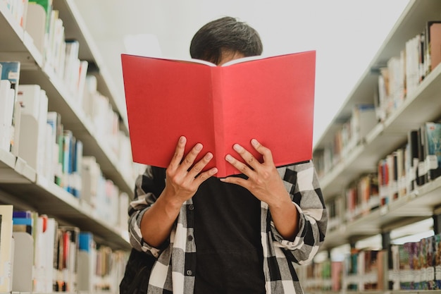 Retrato de un joven estudiante de estilo informal leyendo un libro y cubriéndose la cara con una estantería