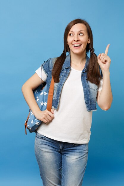 Retrato de joven estudiante hermosa divertida en ropa de mezclilla con mochila apuntando con el dedo índice hacia arriba mirando a un lado aislado sobre fondo azul. Educación en la universidad. Copie el espacio para publicidad.