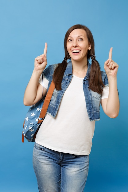 Retrato de joven estudiante mujer atractiva alegre en ropa de mezclilla con mochila mirando hacia arriba con el dedo índice aislado sobre fondo azul. Educación en la escuela secundaria. Copie el espacio para publicidad.