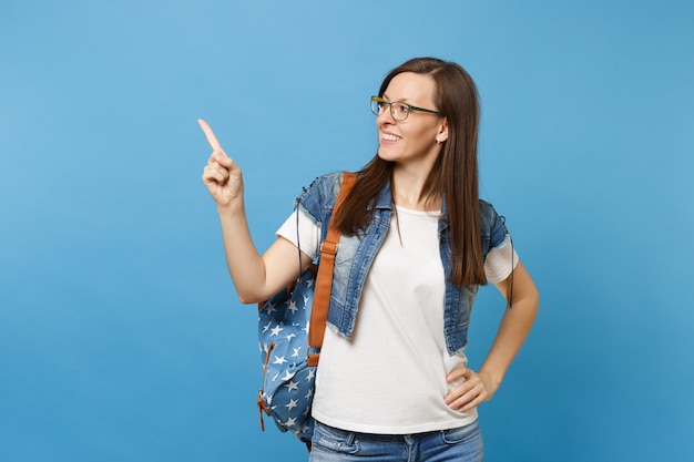 Retrato de joven estudiante mujer bonita en gafas de ropa de mezclilla con mochila mirar a un lado señalar con el dedo en el espacio de la copia aislado sobre fondo azul. Educación en el concepto de colegio universitario de secundaria.