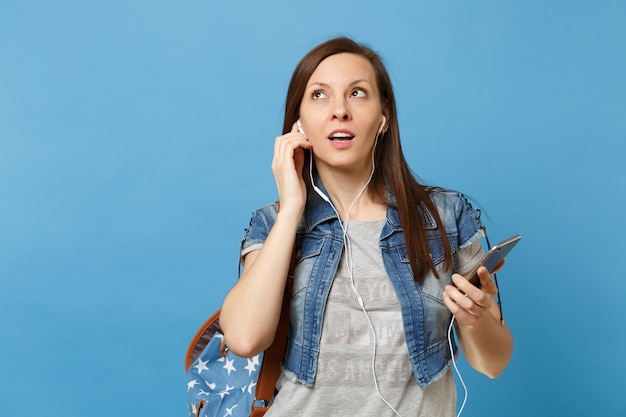 Retrato de joven estudiante mujer curiosa con mochila y auriculares escuchando música sosteniendo teléfono móvil mirando hacia arriba aislado sobre fondo azul. Educación en la universidad. Copie el espacio para publicidad.