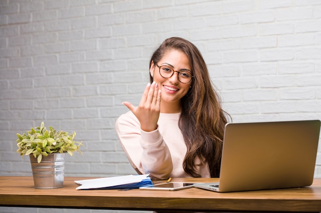 Retrato de joven estudiante mujer latina sentada en su escritorio invitando a venir