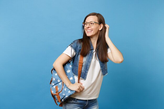 Retrato de joven estudiante mujer riendo con mochila con gafas mirando a un lado tocando corregir su peinado aislado sobre fondo azul. Educación en el concepto de colegio universitario de secundaria.