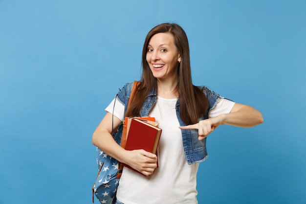 Retrato de joven estudiante mujer sonriente emocionada con mochila sosteniendo libros escolares apuntando con el dedo índice en el espacio de la copia aislado sobre fondo azul. Educación en el concepto de colegio universitario de secundaria.