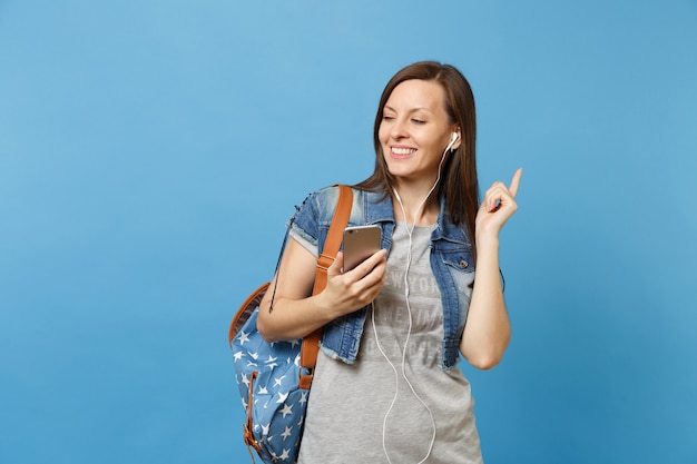 Retrato de joven estudiante mujer sonriente relajada con mochila y auriculares escuchando música sosteniendo teléfono móvil aislado sobre fondo azul. Educación en la escuela secundaria. Copie el espacio para publicidad.