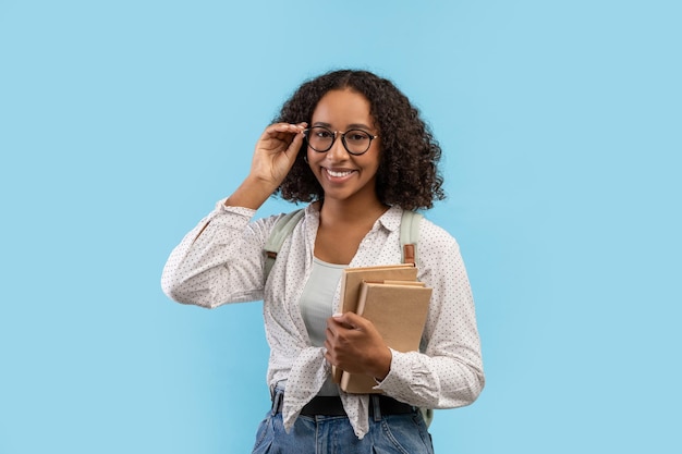 Retrato de una joven estudiante negra con libros tocando gafas y sonriendo a la cámara sobre azul