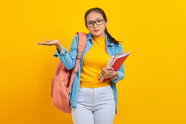 Retrato de una joven estudiante universitaria asiática confundida con ropa de mezclilla con mochila sosteniendo un cuaderno y señalando el espacio de copia con la mano aislada en el fondo amarillo